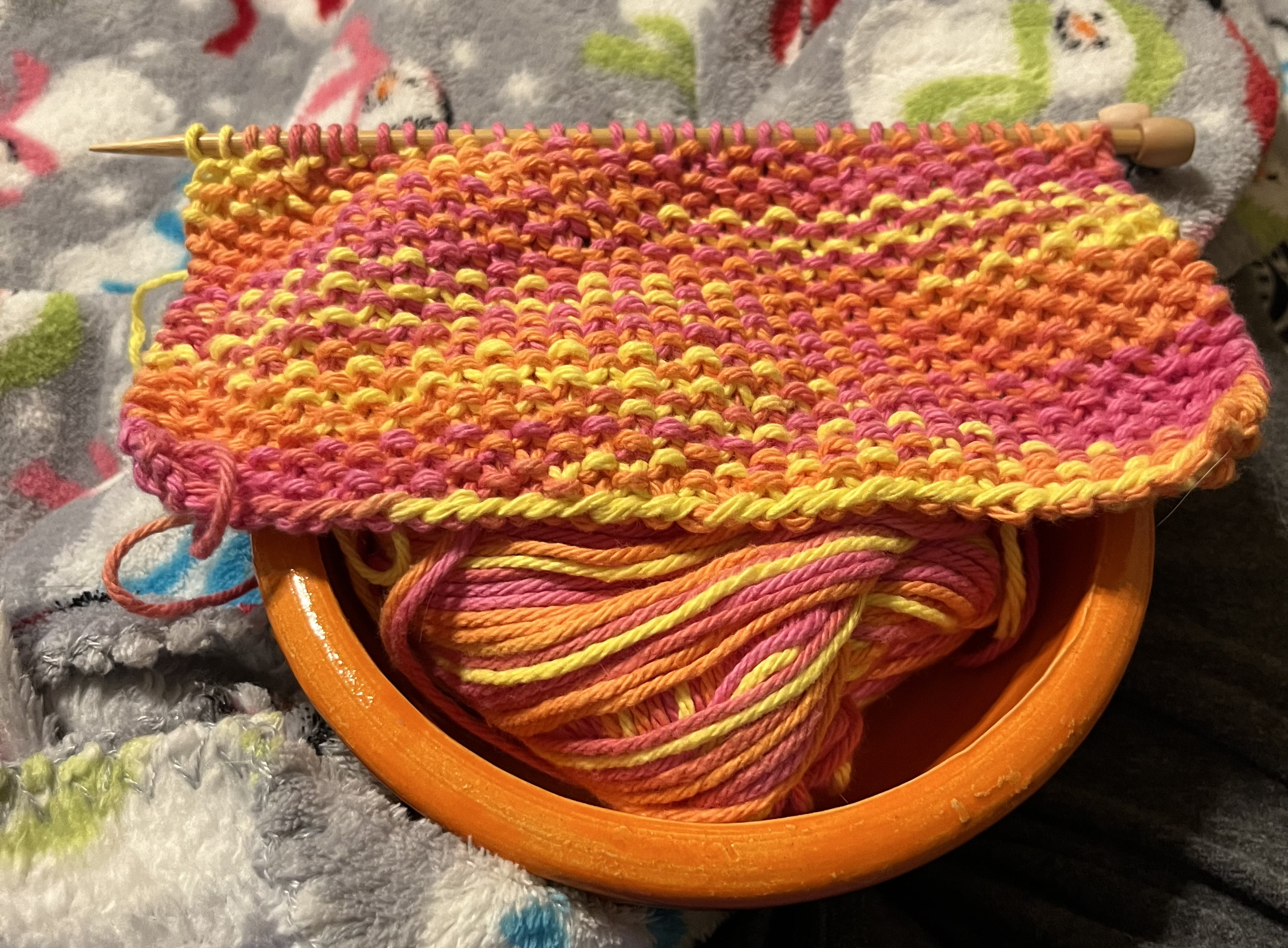 A partly completed knitted dishcloth in seed stitch.  It is pink, orange, and yellow, and sits on top of an orange bowl.